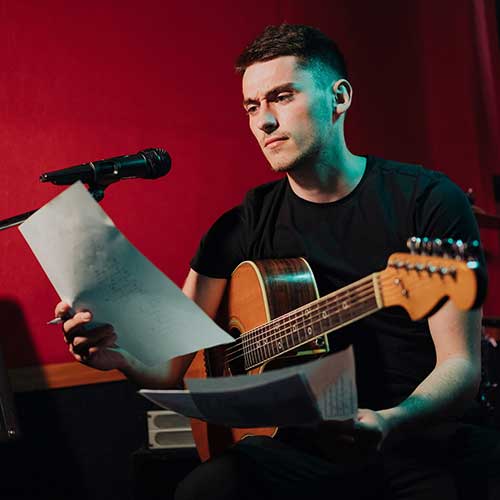 A songwriter, with guitar in lap and pen in hand, looking at a lyric sheet