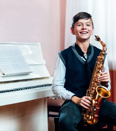Cheerful student boy sitting with saxophone before music lesson on piano background